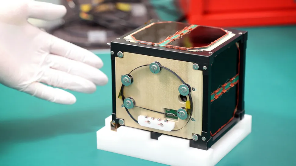 a small cubical satellite sits on a blue-green tabletop, with a white-gloved hand next to it for perspective